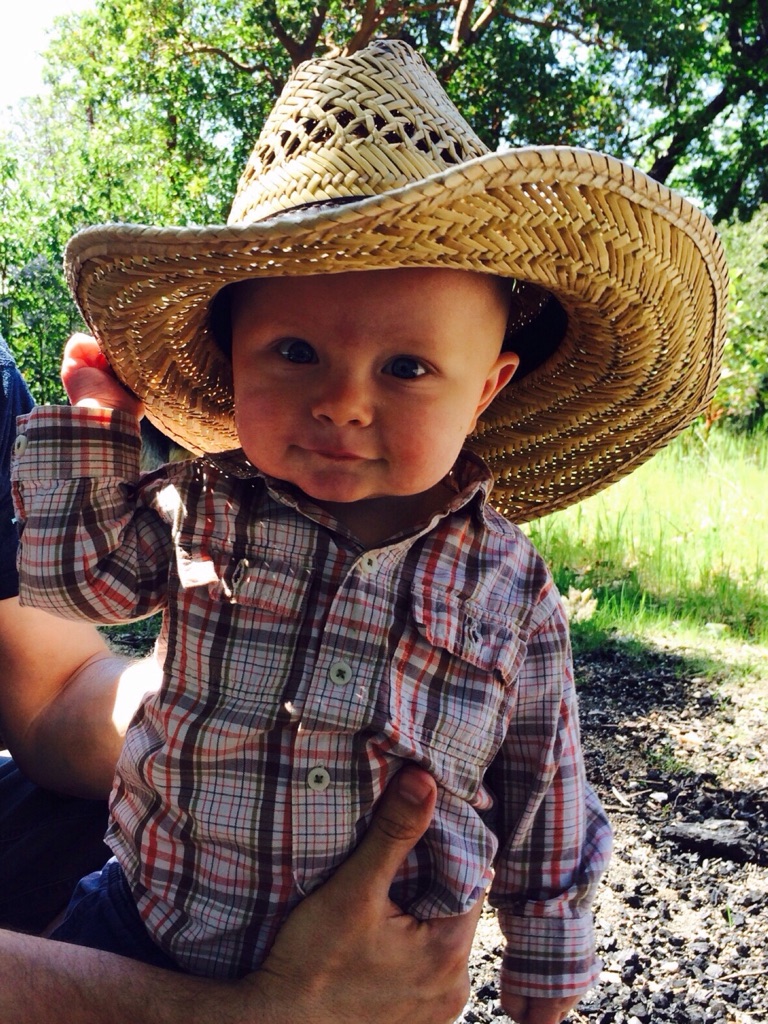 9-month-old Eli wearing his father's cowboy hat and a plaid button-up shirt.